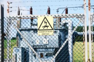 A fenced-off electrical substation with a yellow warning sign reading “Danger of Death – Keep Out,” surrounded by barbed wire for safety.