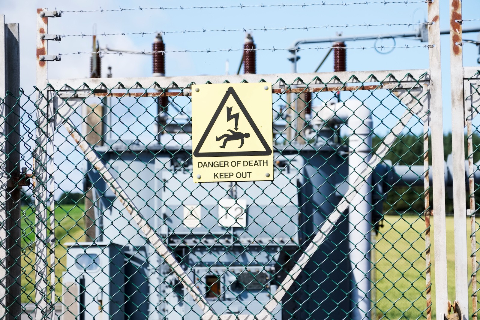 A fenced-off electrical substation with a yellow warning sign reading “Danger of Death – Keep Out,” surrounded by barbed wire for safety.
